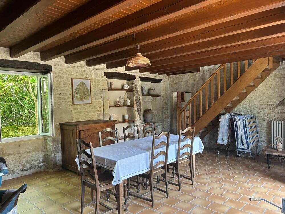 Vue d'ensemble du salon du Gîte Pampas au Domaine du Vieux Lavoir, prise d'un autre angle, révélant l'espace repas avec sa table et ses chaises, ainsi que l'escalier menant au premier étage. Cet aménagement complet assure un confort optimal pour les groupes et familles séjournant sur place, idéal pour les mariages et grandes réceptions dans les Deux-Sèvres. Un lieu de vie fonctionnel et chaleureux pour vos invités.