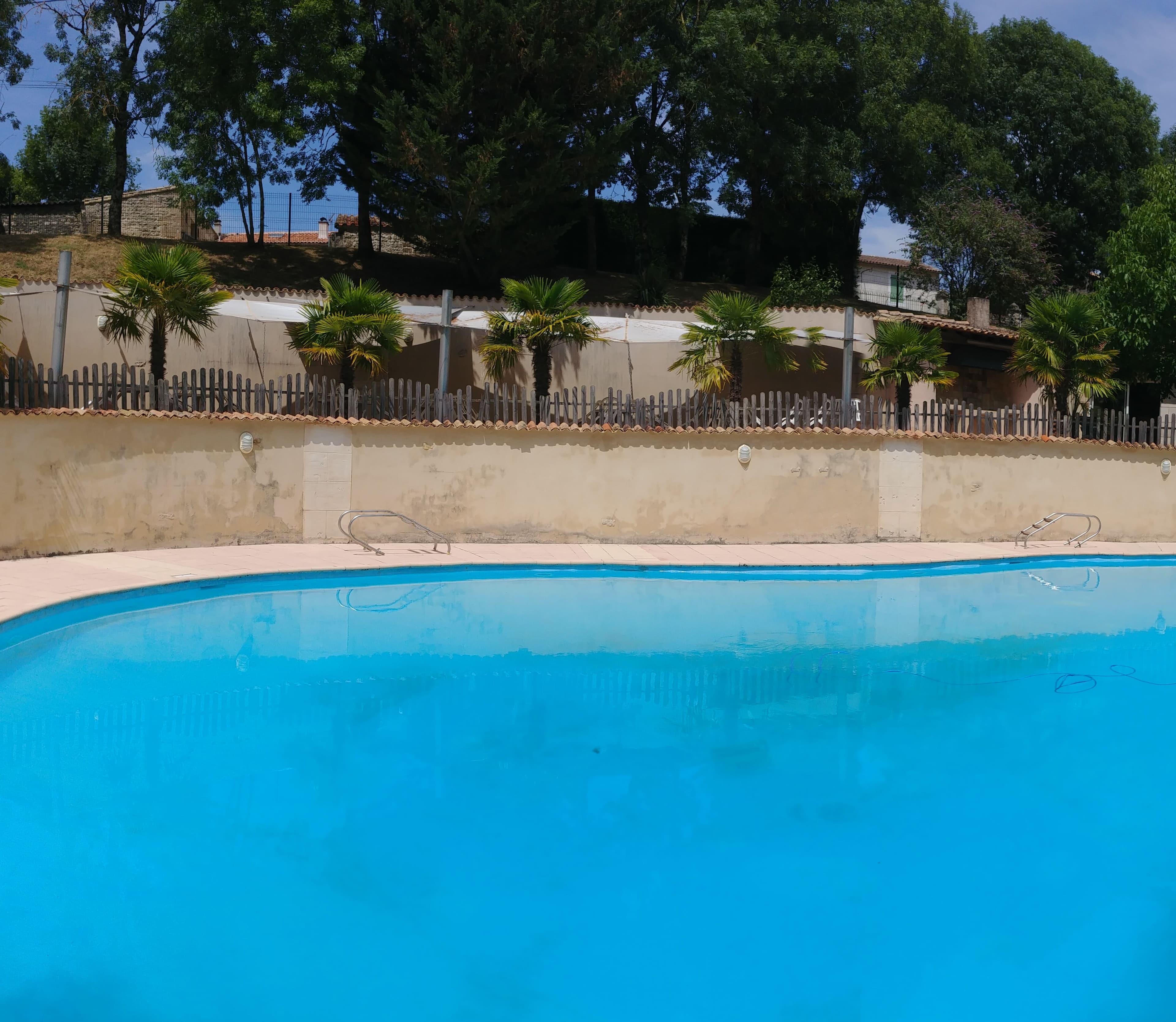 La magnifique piscine du Domaine du Vieux Lavoir au premier plan, avec en arrière-plan la terrasse ombragée par des voiles élégantes, qui surplombe la piscine. Cet aménagement offre un espace idyllique pour la détente et le rafraîchissement des invités de vos mariages et grandes réceptions dans les Deux-Sèvres, permettant de profiter pleinement du cadre ensoleillé tout en bénéficiant d'un abri confortable et d'une vue agréable.
